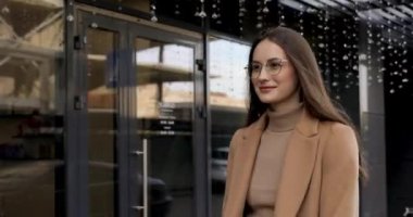 Uzun kahverengi gözlüklü ve bej ceketli şık bir kadın sokakta yürüyor. Mutlu genç bayan açık havada temiz sonbahar havasının tadını çıkarıyor..