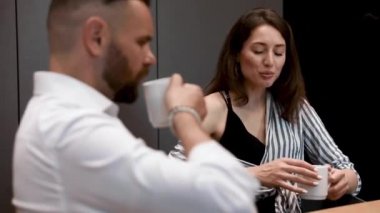 Çizgili gömlekli çekici bir bayan bir fincan kahveyle masaya oturuyor ve kahvaltıda sakallı kocasıyla konuşuyor. Güzel evli çift sabah birlikte mutfakta vakit geçiriyorlar..