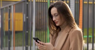 Göz giysisi ve bej ceketli çekici bir esmer, temiz havada akıllı telefon ekranına bakarken mutlu oluyor. Şehir yaşam tarzı ve modern teknoloji kavramı.