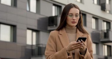 Gözlüklü ve bej ceketli ciddi güzel bir kadın karanlık binanın yanında dururken akıllı telefon kullanıyor. Genç esmer, internette geziyor..