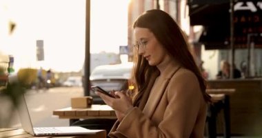 Gözlüklü ve sonbahar montu giyen gülümseyen genç bir kadının açık dizüstü bilgisayarlı ve elinde akıllı telefonla terasta oturması. İnsanlar ve modern teknoloji kavramı.