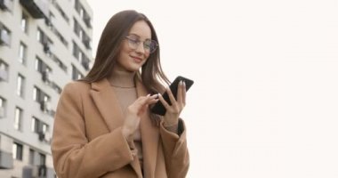Uzun kahverengi saçlı, gülümseyen güzel bayan şehir merkezinde modern akıllı telefon kullanıyor. Gözlük takan ve bej rengi sonbahar ceketi giyen genç bir kadın. Yaşam tarzı kavramı.