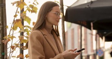 Gözlüklü ve bej ceketli güzel bir bayanın sokakta dikilip sosyal ağ için modern akıllı telefonu kullanışının yan görüntüsü. İnsanlar ve teknoloji kavramı.