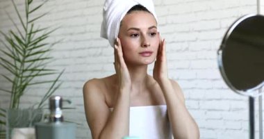 Kafasında beyaz havlu olan güzel bir kadın parlak banyoda otururken saf yüzüne parmaklarıyla nazikçe dokunuyor. Genç bayan evde yüz masajı yapıyor..