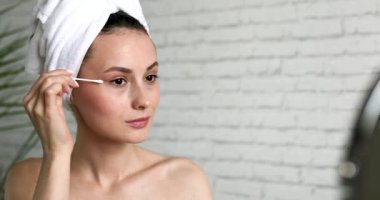 Göz altı makyajı için pamuklu bez kullanarak banyo havlusuna sarılmış çekici bir kadın. Banyoda aynaya bakan çıplak omuzlu mutlu bir esmer..
