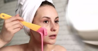 Çıplak omuzlu rahat genç bayan sarı aplikatörlü pembe jel maske takıyor. Kafasında havlu olan hoş bir kadın banyoda spa tedavisi uyguluyor..