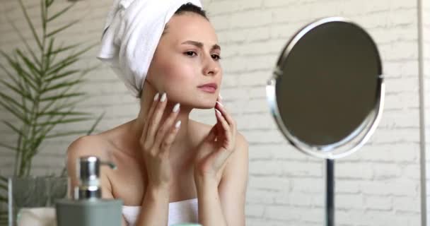 Junge Frau im Badetuch blickt in den Spiegel und sieht wegen ihres Hautbildes enttäuscht aus. Natürliches Schönheitskonzept. Behandlungen zu Hause. — Stockvideo