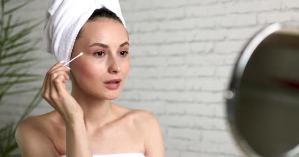 Encantadora joven con toalla de baño en la cabeza limpiando la piel bajo los ojos con hisopo de algodón. Hermosa mujer disfrutando de rituales matutinos en casa. — Vídeos de Stock