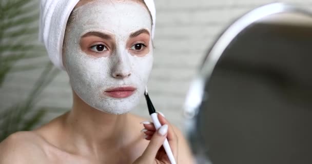 Szczęśliwa młoda kobieta stosująca nawilżającą białą maskę ze szczotką w łazience. Przystojna pani z naturalnym pięknem uśmiechnięta i patrząca w kamerę. — Wideo stockowe