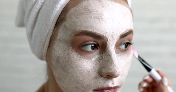 Közelkép egy csinos nőről, fehér törölközőbe csomagolva, maszkkal az arcán, kis ecsettel. Házi készítésű bőrápolás. Reggel a fürdőszobában.. — Stock videók