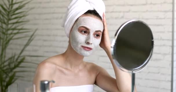 Молодая женщина позирует на камеру в белой косметической маске на лице. Очаровательная брюнетка с голыми плечами сидит в ванной и смотрит на круглые зеркала. — стоковое видео
