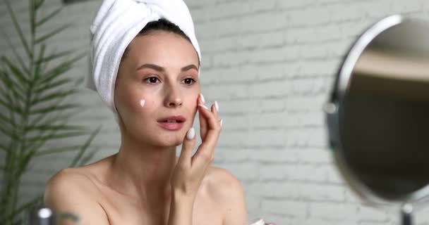 Mujer atractiva que aplica crema hidratante en la cara después de la ducha de la mañana. Jovencita mirando el espejo mientras está sentada en el baño luminoso. — Vídeo de stock