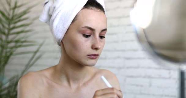 Aantrekkelijke jonge brunette aanbrengen van wit masker op het gezicht met cosmetische borstel. Ontspannen vrouw gewikkeld in handdoek na de douche. Ochtendroutine thuis. — Stockvideo