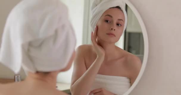 Ελκυστική νεαρή γυναίκα τυλιγμένη σε πετσέτα μπάνιου αγγίζοντας απαλά το πρόσωπό της με τα δάχτυλα. Η όμορφη κυρία εκτιμά την κατάσταση του δέρματός της. Καλλυντικά και διαδικασίες. — Αρχείο Βίντεο