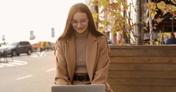 Mooie jonge vrouw in bril en warme jas met laptop terwijl ze op een houten bank zit. Herfst sfeer rond. Begrip werk op afstand. — Stockvideo