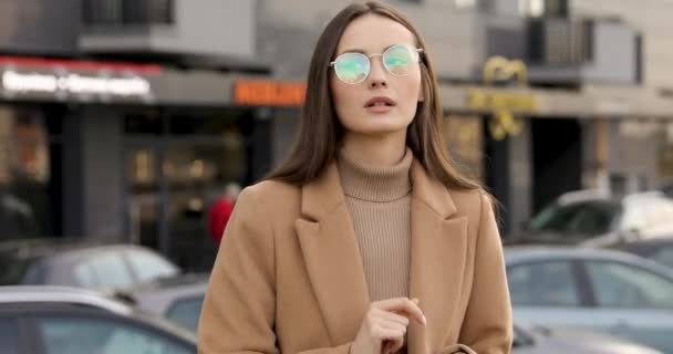 Ragazza premurosa che indossa cappotto beige e occhiali in piedi sulla strada, tenendo smartphone e guardarsi intorno. Concetto di persone e tecnologia. — Video Stock