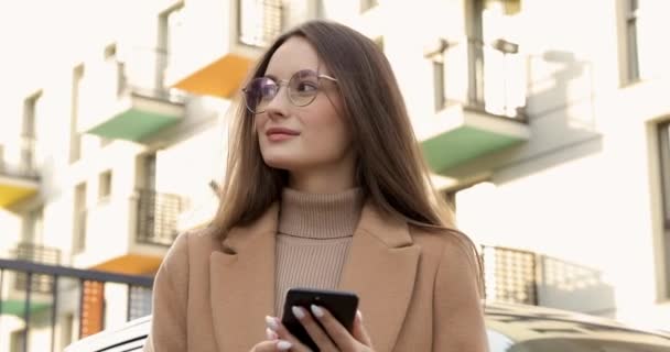 Piacevole giovane femmina con bellezza naturale in cappotto beige e occhiali utilizzando smartphone all'aperto. Concetto di stile di vita cittadino, persone e tecnologia moderna. — Video Stock