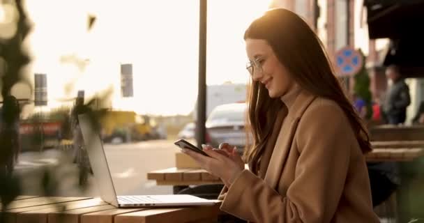 Glad ung brunett i glasögon och beige rock sitter vid bordet och använder smartphone och laptop. Vacker kvinna som arbetar med moderna prylar på öppen terrass. — Stockvideo