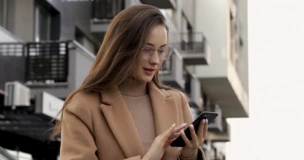 Encantadora joven morena en gafas y abrigo beige respondiendo correos electrónicos en el teléfono inteligente mientras está de pie en la calle. Concepto de conveniencia y tecnología moderna. — Vídeos de Stock