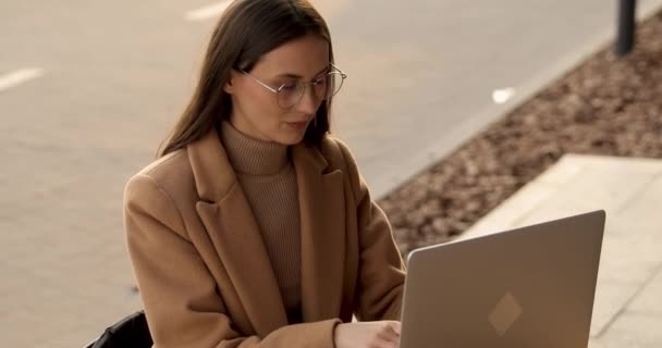 Encantadora freelancer femenina en abrigo beige y gafas que utiliza portátil inalámbrico para trabajar en el aire fresco. Feliz joven sentada en la acogedora cafetería y trabajando remotamente en la computadora. — Vídeos de Stock