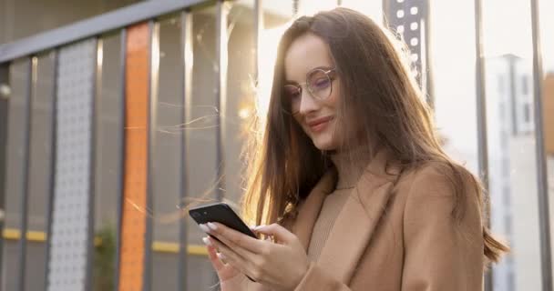 Porträt einer hübschen jungen Frau mit langen braunen Haaren, die mit einem modernen Smartphone in der Hand vor der Kamera posiert. Charmante Frau im beigen Mantel verbringt ihre Freizeit an der frischen Luft an sonnigen Herbsttagen. — Stockvideo