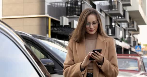 Jeune femme confiante aux longs cheveux bruns s'appuyant sur la voiture dans la rue et utilisant un smartphone moderne. Belle femme portant des lunettes et manteau d'automne beige. — Video