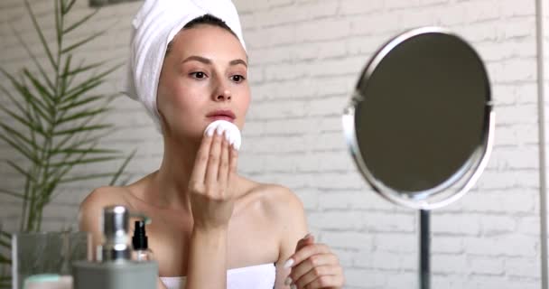 Mujer sana y joven con el pelo envuelto en toalla de baño cara de limpieza con loción y almohadilla de algodón. Tratamiento facial matutino en casa. — Vídeo de stock