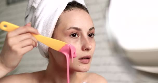 Jeune femme détendue aux épaules nues mettant un masque rose semblable à un gel sur le visage avec applicateur jaune. Agréable femme avec serviette sur la tête faisant des soins spa à la salle de bain. — Video