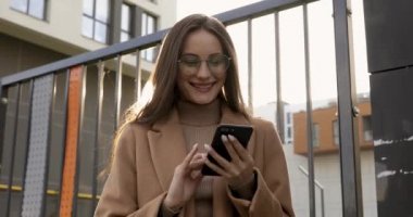 Gözlüklü ve ceketli neşeli genç bir kadın sokakta dikilirken akıllı telefondan mesaj yazıyor. Kahverengi saçlı güzel bir kadın arkadaşlarıyla internet üzerinden iletişim kuruyor..
