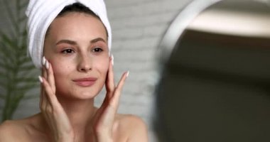 Çıplak omuzlu güzel bir kadın sabah duşundan sonra yüz kremi sürüyor. Kafasında havlu olan mutlu genç bayan evde nemlendirici cilt.