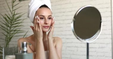 Çıplak omuzlu, sabah duşundan sonra yanaklarına masaj yapan hoş bir genç bayan. Mutlu esmer kadın aynaya bakıyor ve evde cilt bakımı yapıyor..