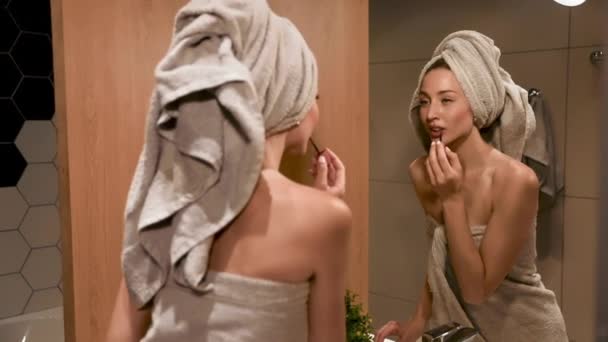 Жінка в рушничку дивиться на дзеркало і наносить помаду — стокове відео