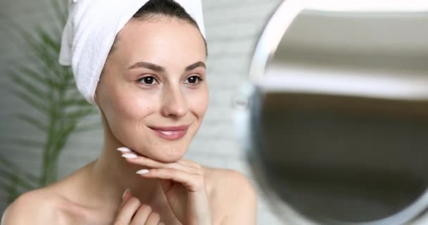 Angenehme junge Frau im Badetuch, die in den Spiegel schaut und ihre frische, reine Haut genießt. Naturschönheit. Morgens Zeit für sich selbst. — Stockvideo