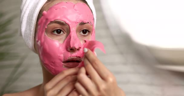 Mujer joven feliz con toalla en la cabeza quitándose la máscara de alginato rosa de la cara mientras mira el espejo redondo. Señora atractiva usando cosméticos profesionales regularmente en casa. — Vídeos de Stock