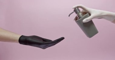 Bir elin beyaz eldivende, elinde sıvı sabunla siyah eldivene basıyorsun. Pembe stüdyo arka planında izole edilmiş. Hijyen ve koruma kavramı.