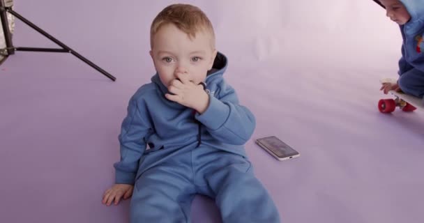 Niño pequeño sentado en el suelo con el móvil moderno, mientras que otro niño montando monopatín cerca. Dos niños en ropa casual posando en estudio con fondo violeta. — Vídeos de Stock