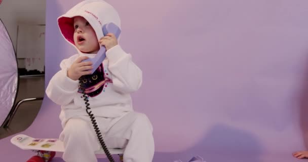 후디에 앉아 스케이트 보드에 앉아 빈티지 전화기로 이야기하는 장난스럽고 귀여운 아기. 손을 잡고 있는 예쁜 아기, 집안에서 몸짓을 하고 미소짓는 모습. — 비디오
