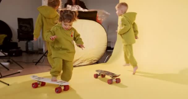 Szczęśliwe dzieci w różnym wieku ubrane w podobny kombinezon, bawiące się deskorolkami w studio z żółtym tłem. Szczęśliwe dzieciństwo i zabawny czas. — Wideo stockowe