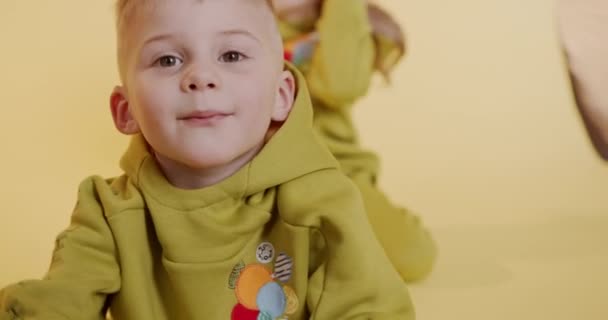 Playful cute boy in hoodie showing his tongue on camera while female friends playing behind. Yellow studio background. Happy kids enjoying time together. — Stock Video