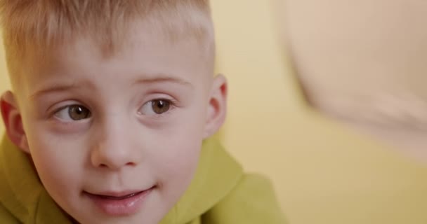 Aranyos kisfiú portréja, világos hajjal, aki trükköket játszik a kamerában. Homályos háttér boldog gyerek játszik a padlón. — Stock videók