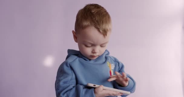 Portrait de joli garçon en sweat à capuche bleu jouant à des jeux sur smartphone moderne. Isolé sur fond de studio violet. Concept de technologie et d'enfance. — Video
