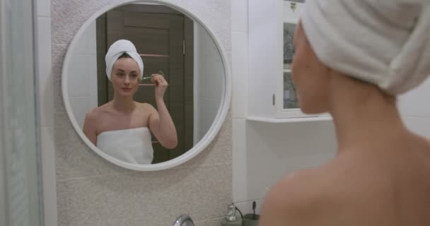 흰 수건을 쓴 아름다운 여성이 눈 밑의 피부 상태를 관리하고 있습니다. 아름다운 자연 그대로의 여성이 화장실에 서서 거울을 보는 모습. — 비디오