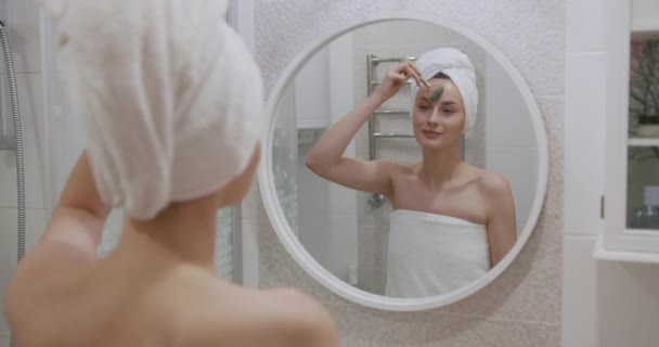 Aantrekkelijke vrouw gewikkeld in witte handdoek met behulp van cosmetische roller voor het heffen van massage op het gezicht. Mooie vrouw op zoek naar spiegel terwijl staan op de badkamer. — Stockvideo