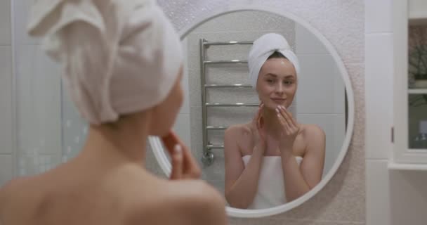 Jovem com toalha de banho na cabeça olhando para o espelho e desfrutando de sua pele perfeita e clara. Conceito de tratamento de beleza e procedimentos. — Vídeo de Stock