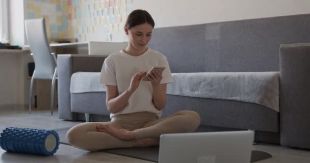 Gelukkige jonge dame in activewear zittend op yoga mat en met behulp van smartphone. Massage roller en laptop liggen op de vloer. Concept van moderne gadgets en thuistraining. — Stockvideo