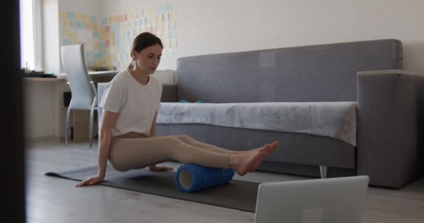 Mujer activa usando rodillo de espuma para la relajación de las piernas después del entrenamiento en casa. Mujer viendo tutorial en línea en el portátil moderno. Equipo deportivo para eliminar el dolor en los músculos. — Vídeo de stock