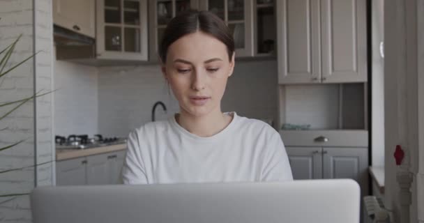 Schöne junge Frau in Freizeitkleidung sitzt am Tisch und arbeitet an einem modernen Laptop. Freiberuflerin nutzt tragbaren Computer für Fernarbeit zu Hause. — Stockvideo