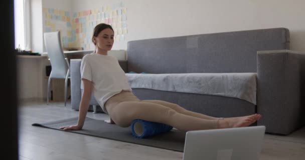 Atrakcyjna kobieta relaksująca nogi na wałku do masażu po porannym treningu. Młoda brunetka w aktywnym ubraniu leżąca na macie do jogi i oglądająca filmy na bezprzewodowym laptopie. — Wideo stockowe