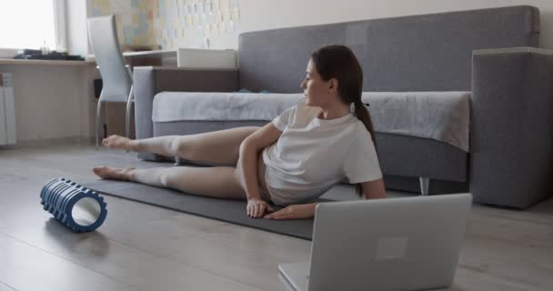 ヨガマットで脚の体操をするスポーツの足やTシャツの若いアクティブな女性。国内ワークアウトのための現代のラップトップを使用して魅力的な女性. — ストック動画