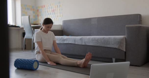 Mujer atractiva sentada en una esterilla de yoga y respirando profundamente para relajarse después de un duro entrenamiento matutino. Atleta femenina usando portátil inalámbrico durante el entrenamiento en casa. — Vídeo de stock